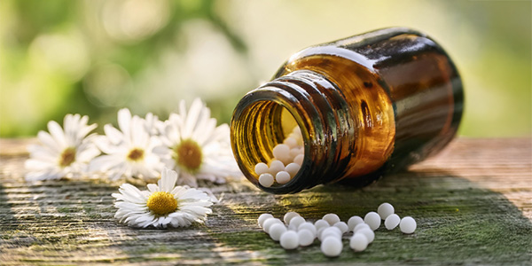 Eine braune Glasflasche liegt auf der Seite auf einem Holztisch; weiße Kügelchen sind herausgefallen. Daneben liegen mehrere weiße Blüten mit gelber Mitte.